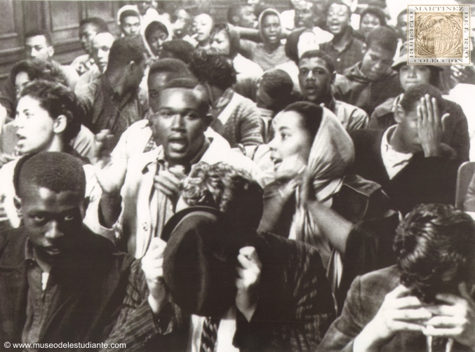 Greensboro. Two white youths arrested in an integration try here hide their faces as some of the mass of Negro students arrested wait booking in Greensboro police headquarters. Over 300 students were arrested in an effort to integrate eating places and moving picture theaters